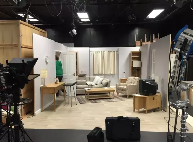 A staged television living-room set surrounded by studio cameras and lighting