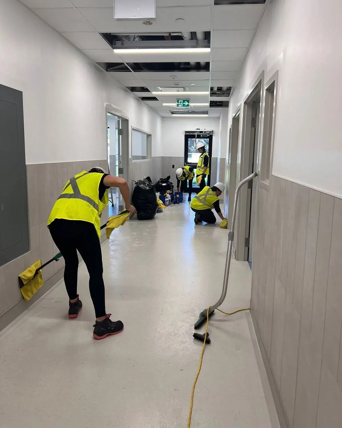A cleaning crew in hi-vis vests and hard hats mopping and vacuuming a corridor during a post-construction cleanup