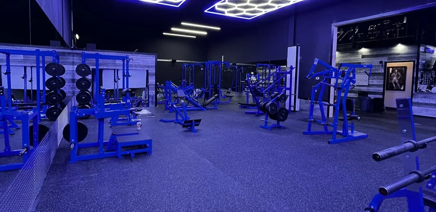 A blue-lit strength-training gym with rows of plate-loaded machines and free weights