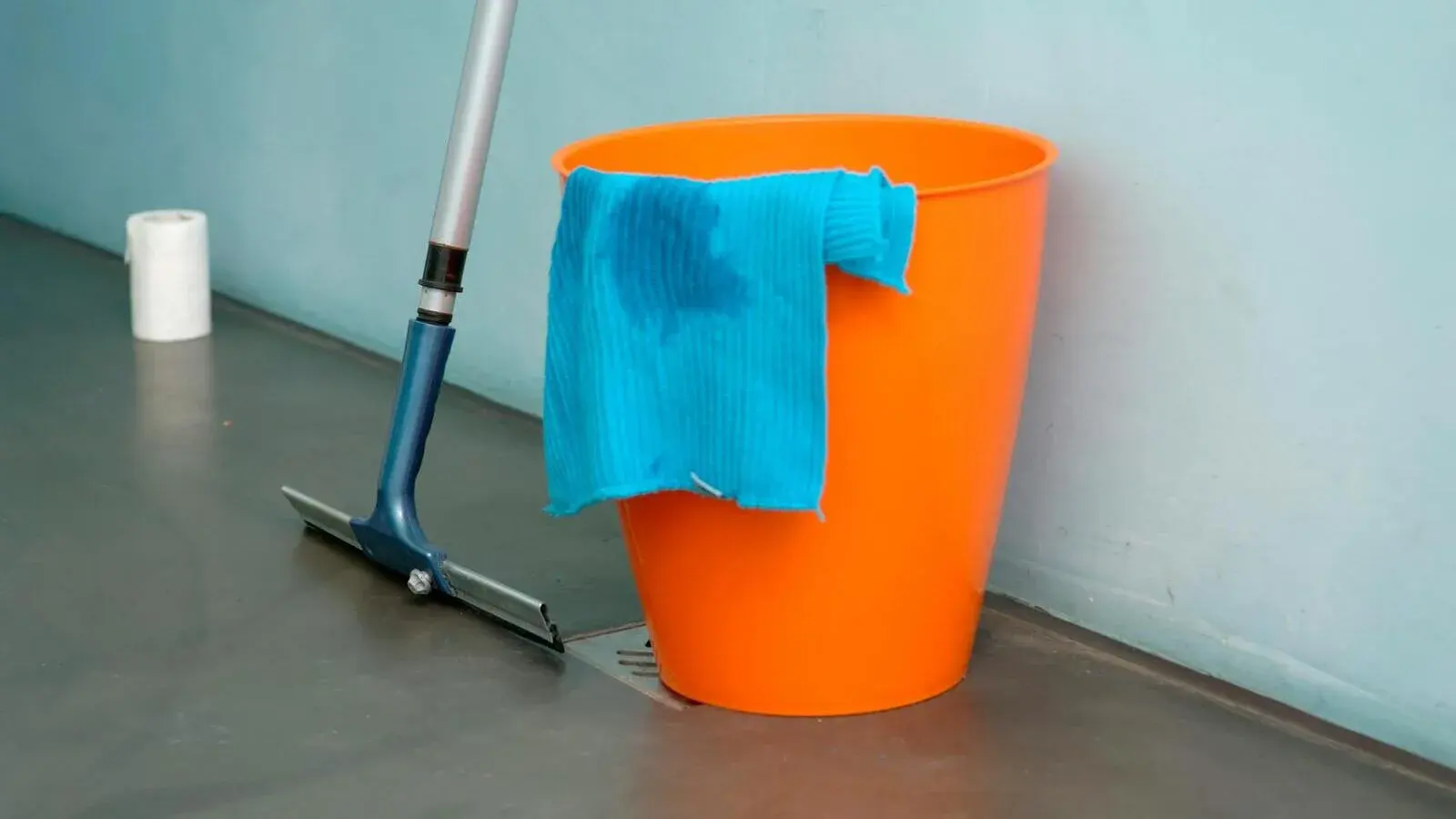 An orange cleaning bucket with a microfiber cloth and a floor squeegee against a wall