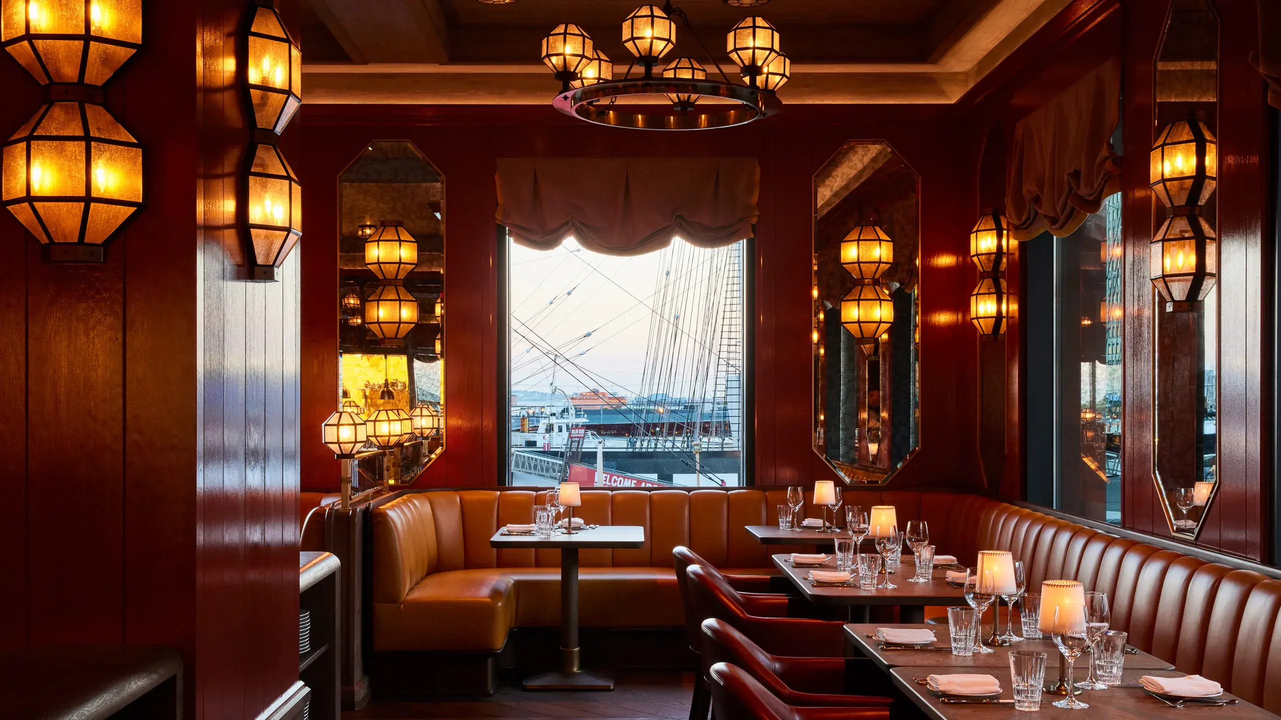 A warmly lit restaurant dining room with leather banquette seating, set tables, and a harbourfront view