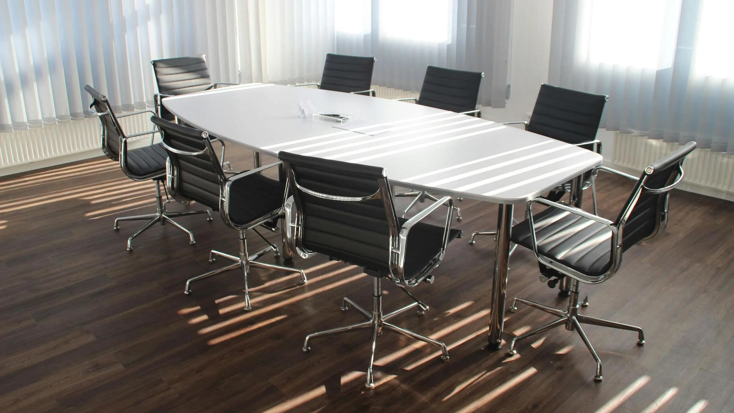 A boardroom with a long conference table and black mesh office chairs, sunlight from tall blinded windows falling across a dark hardwood floor