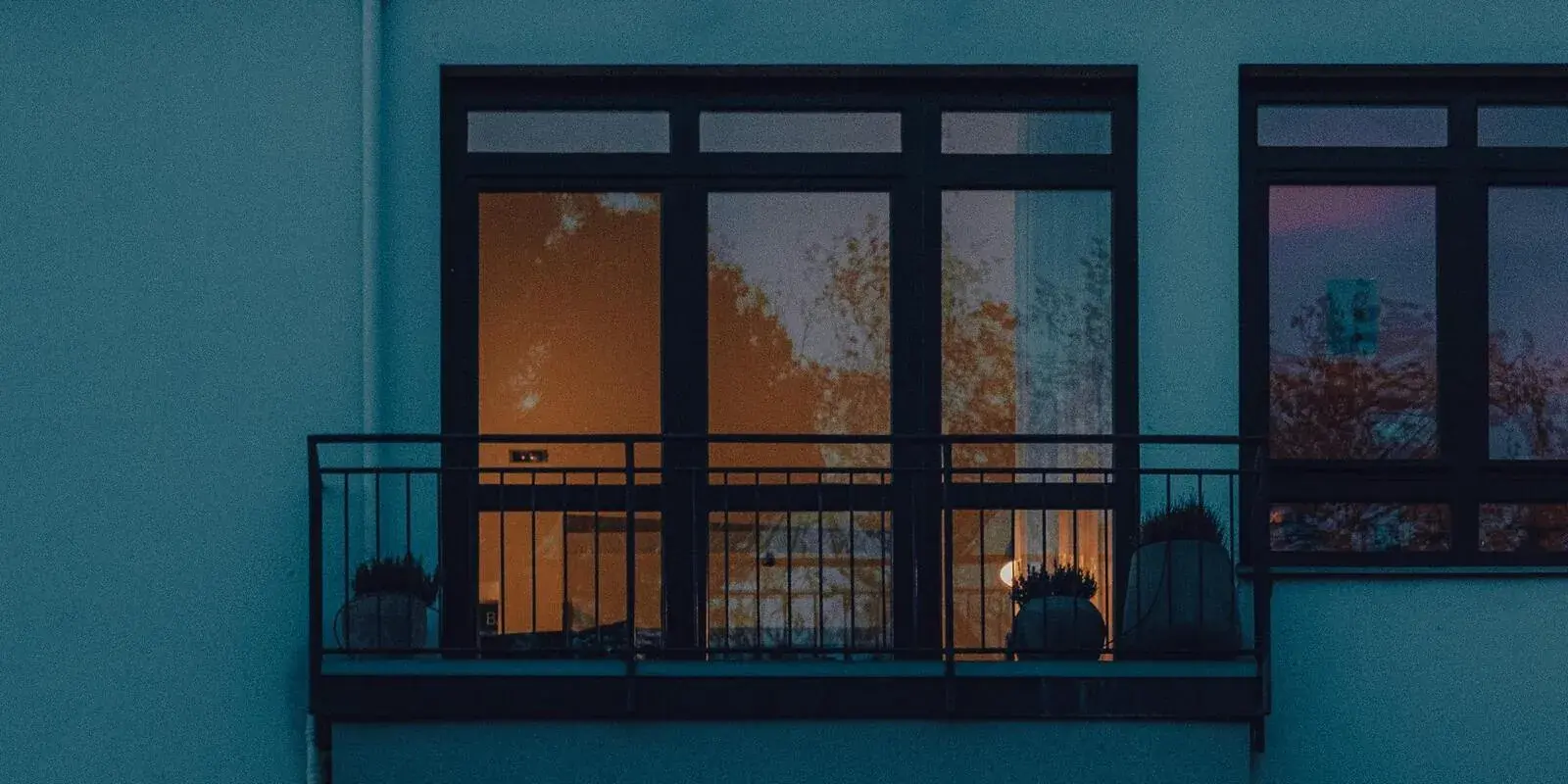 Large apartment windows and a balcony lit warmly at dusk on a multi-tenant residential building