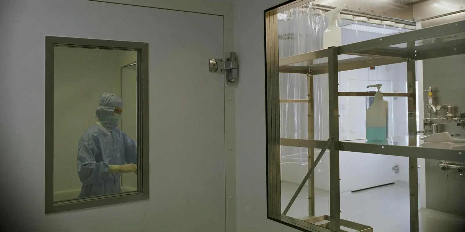 A technician in protective gear seen through the interior glass of a clean clinical lab