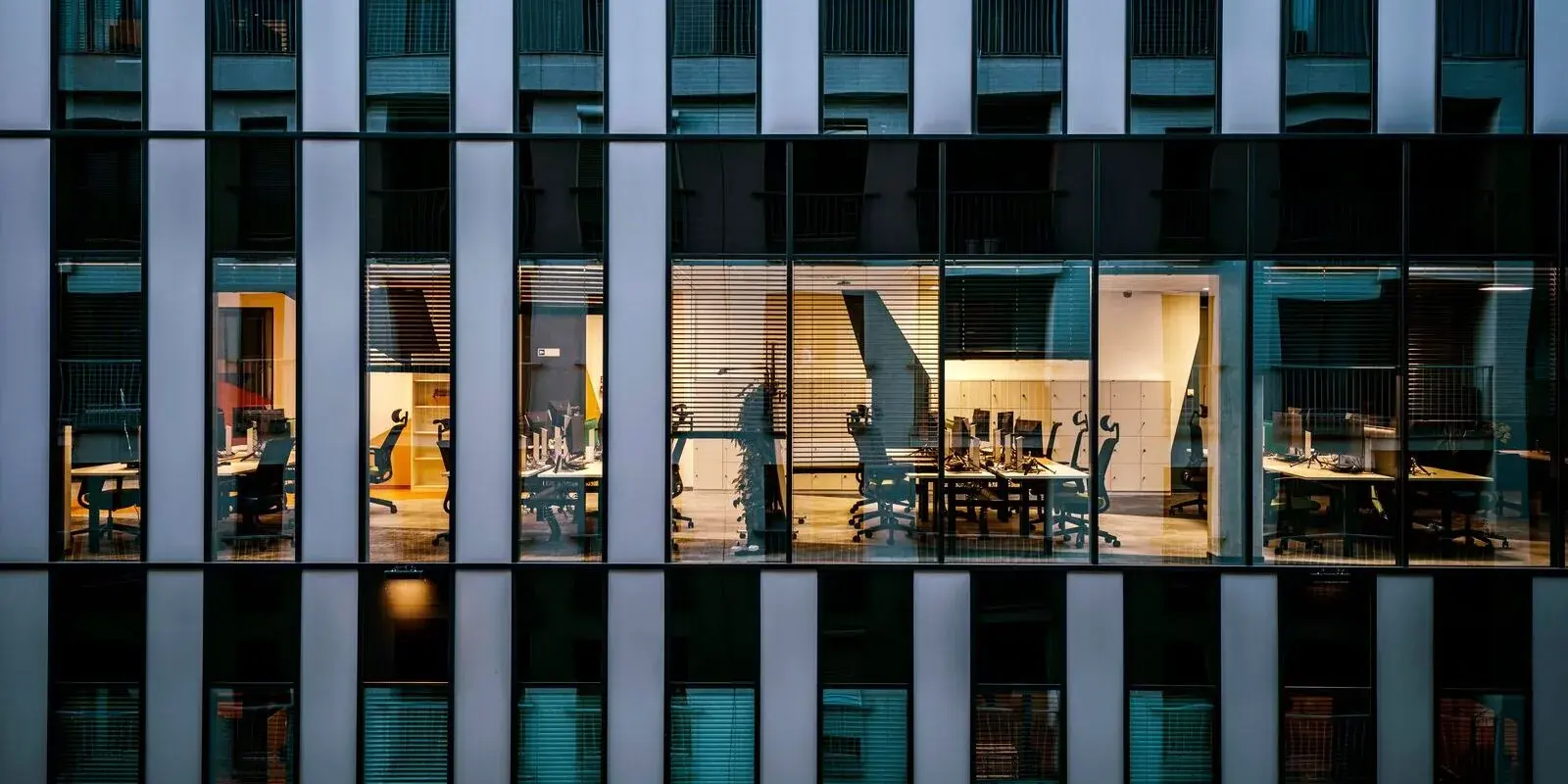 Lit open-plan offices seen through the clean glass facade of a commercial building at dusk